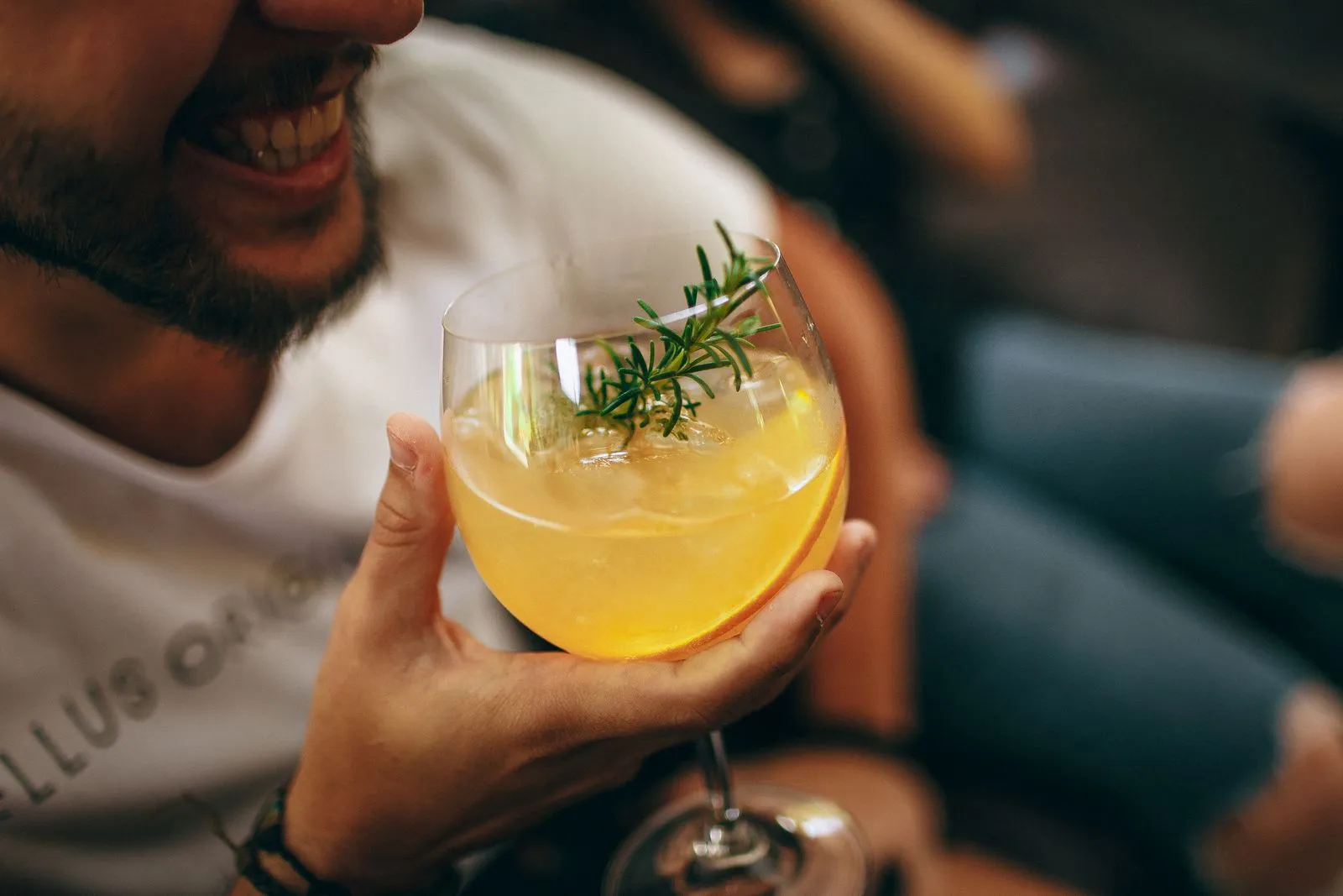 Crop anonymous guy in casual outfit holding glass of cocktail decorated with rosemary while spending time in modern pub during weekend
